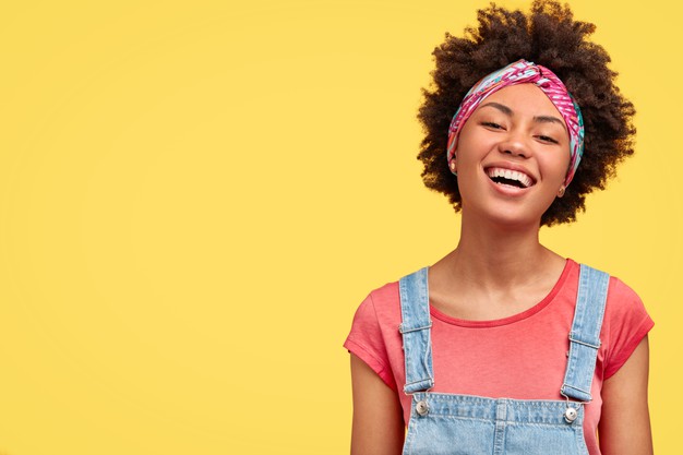 glad young female with dark skin white even teeth laughs positively as sees something funny front wears casual t shirt dungarees isolated yellow wall with blank space aside 273609 15573